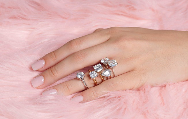 Six engagement rings on display on a woman's hand on a pink background.