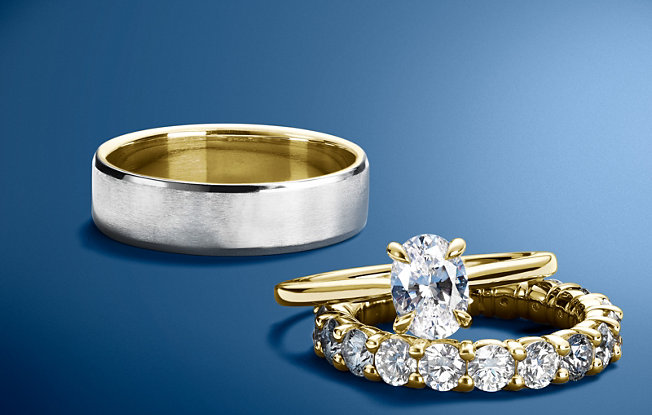 A Blue Nile men's wedding ring and a women's yellow gold engagement ring and wedding band set on a blue background