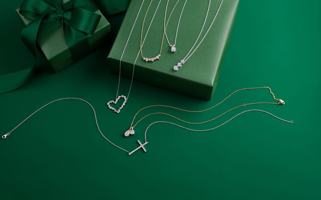 Diamond necklaces in heart, cross and three-stone designs draped around a holiday gift box. 