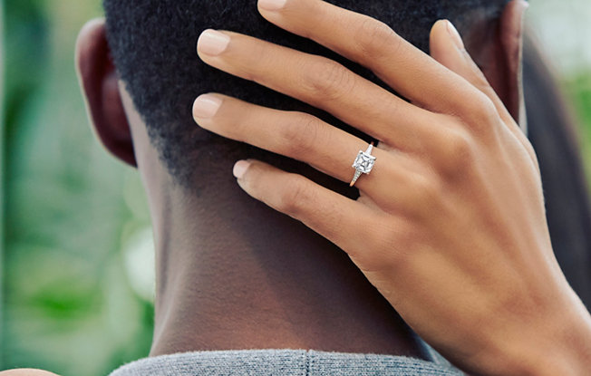 A woman's wearing a cushion cut diamond engagement ring with her hand on the back of a man's head