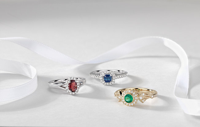 Ruby, topaz and emerald engagement rings of varying cuts on a white background with a white ribbon