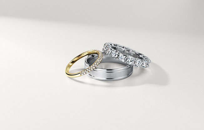A gold wedding band with diamonds, a silver men's wedding ring and a platinum wedding band with diamonds on a white background