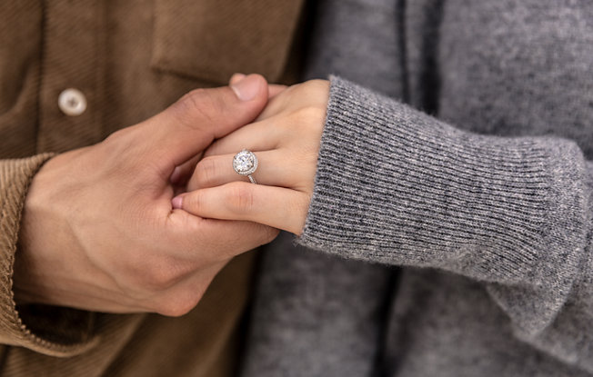 A man wearing a brown jacket and a woman wearing a gray sweater hold hands. The woman is wearing a platinum engagement ring with round center stone and diamond halo