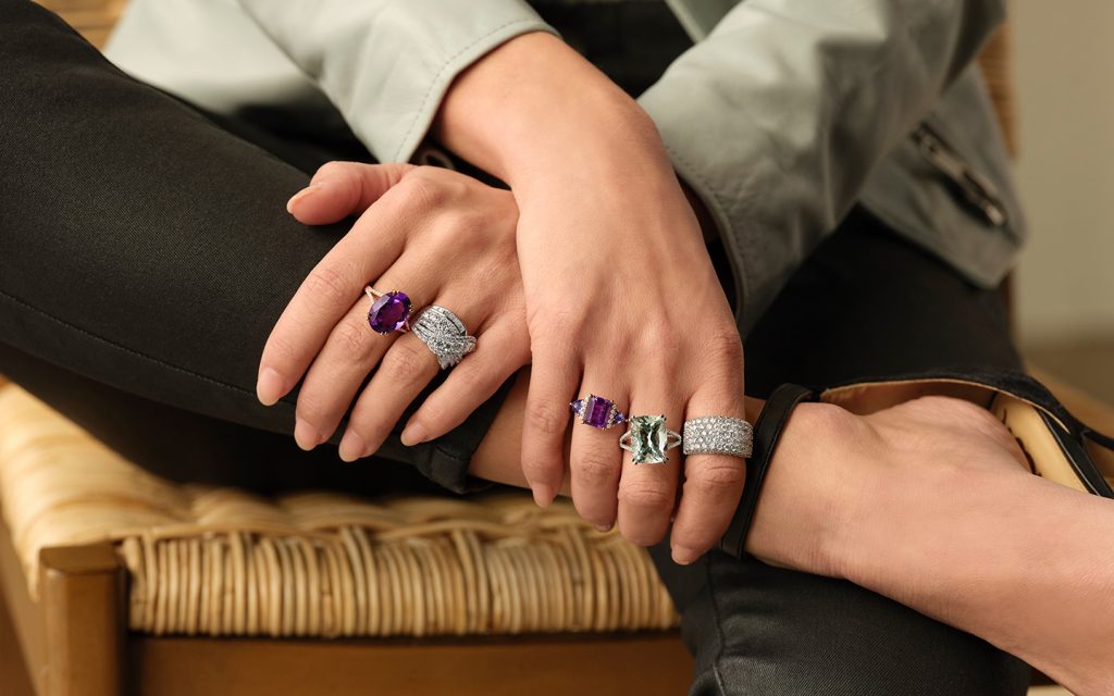 Photo of a woman’s hands; she is wearing five chunky rings with gemstones and diamonds.