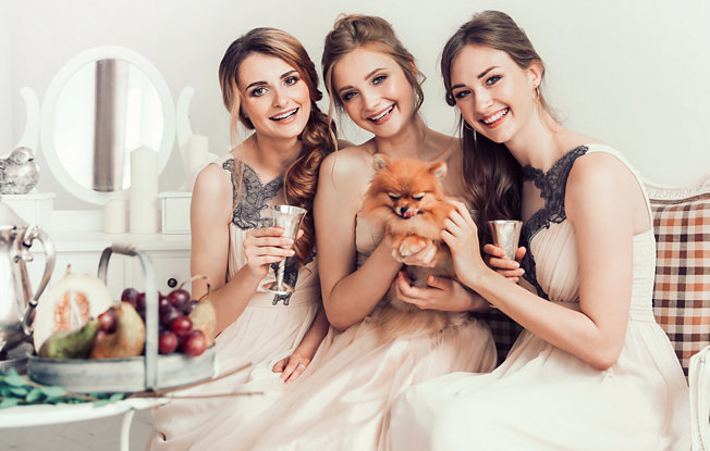 Three girls in bridesmaid dresses cheer while holding a pomeranian puppy