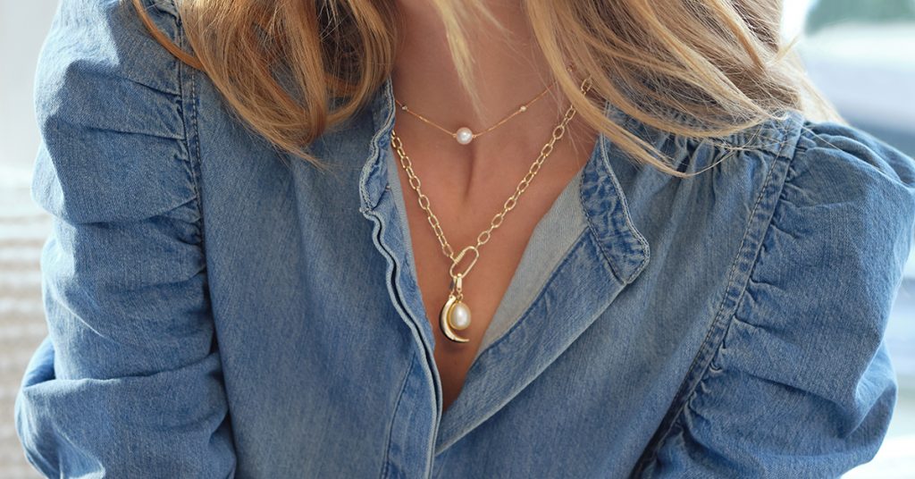 Photo of a woman’s 2 gold necklaces, she is wearing them alongside a denim top.