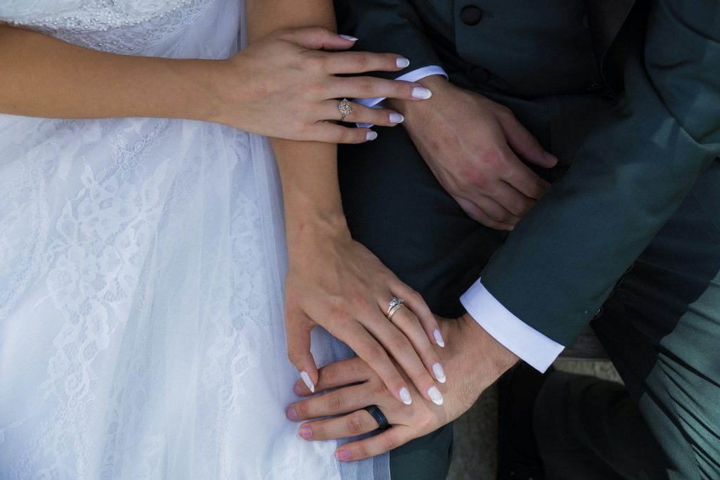 Photo of a bride and groom sitting next to each other with their hands touching, their faces are not visible.