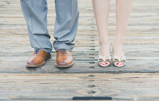 A woman in heels and a man in dress shoes stand on a wooden dock shown from the knees down