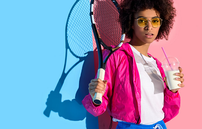 A women holding a tennis racket stands against a pink and blue wall