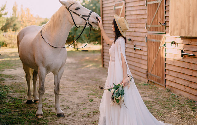 A woman in a wedding dress pats a horse in front of a barn