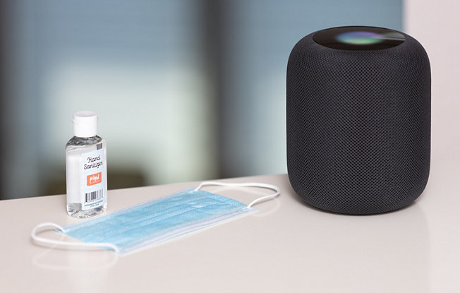 A bottle of hand sanitizer, a blue disposable facemask and a black speaker sit on a wooden table