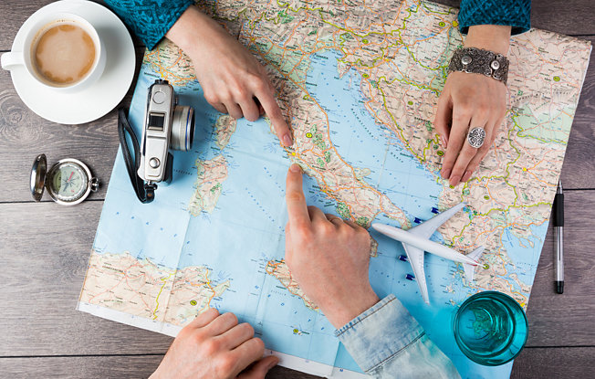 Two hands pointing at a map sitting on a wooden table along with a coffee cup, camera and compass